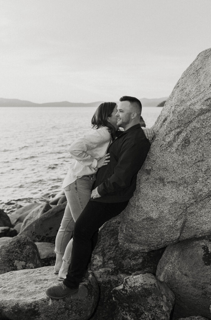 Man leaning on large rock while fiancé whispers in his ear with Lake Tahoe in background at Logan Shoals