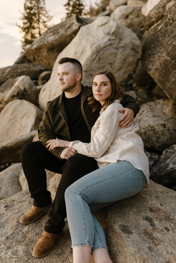 Woman looking at camera while fiancé looks out into distance while engagement couple is sitting on rock at Logan Shoals