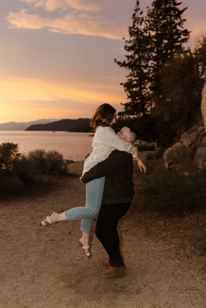 Man picking up fiancé while on a dirt trail with sunset over Lake Tahoe in background at Logan Shoals