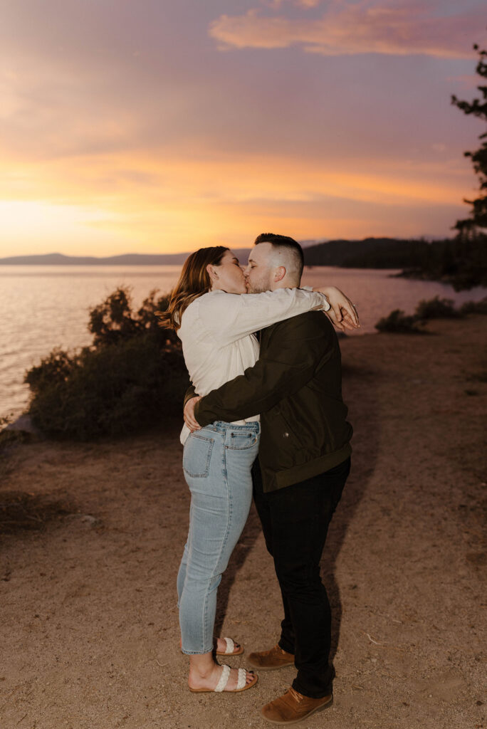 Engagement couple holding and kissing each other while on dirt trail with sunset and Lake Tahoe in background at Logan Shoals