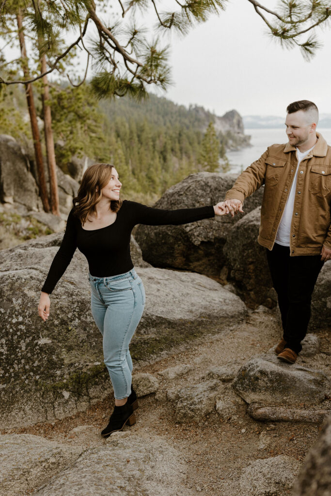 Woman leading fiancé by the hand while looking back at him walking over rocks at Logan Shoals