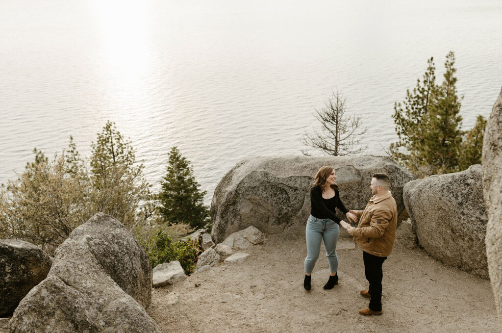 Engagement couple holding hands and smiling at each other while surrounded by large rocks with Lake Tahoe in background at Logan Shoals