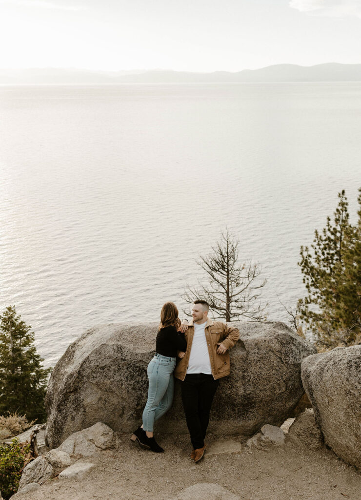 Engagement couple leaning onto large rock together while woman looks out onto Lake Tahoe at Logan Shoals