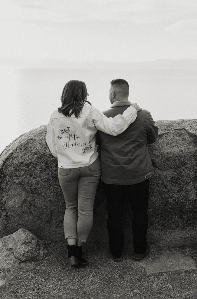Engagement couple standing together looking out at Lake Tahoe with large rock in front of them while woman wears engagement jacket at Logan Shoals