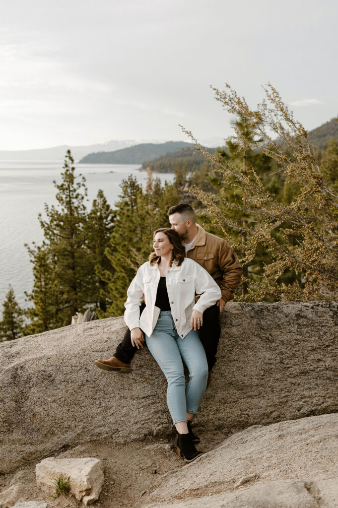 Engagement couple siting on large rock and looking at Lake Tahoe together while man kisses fiancé's head at Logan Shoals