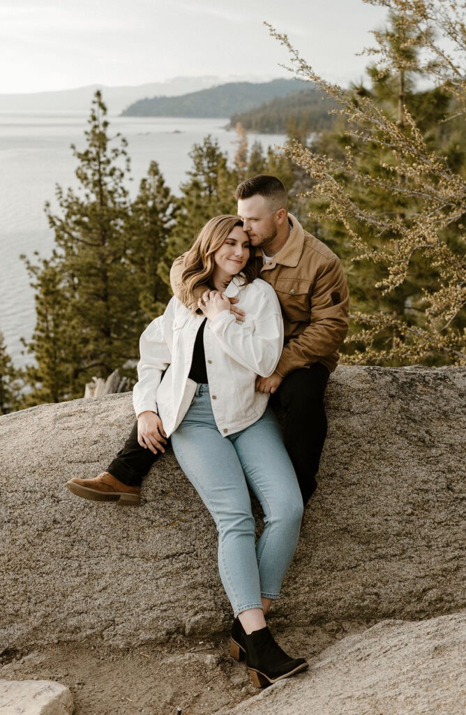 Engagement couple sitting on rock together while holding each other and smiling with pine trees and Lake Tahoe in background at Logan Shoals