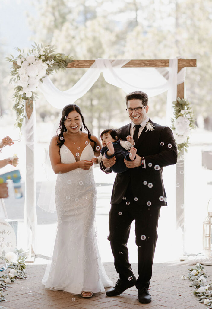 Elopement couple walking down aisle after ceremony while smiling and holding their baby as bubbles blow around them in Lake Tahoe