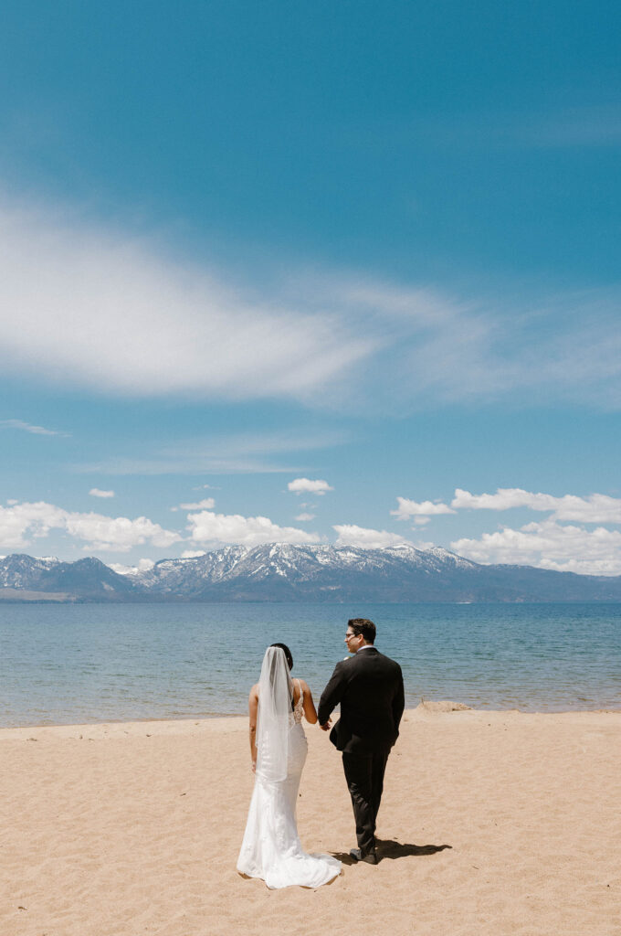 Elopement couple walking down Lake Tahoe beach together in formal attire with mountains in background