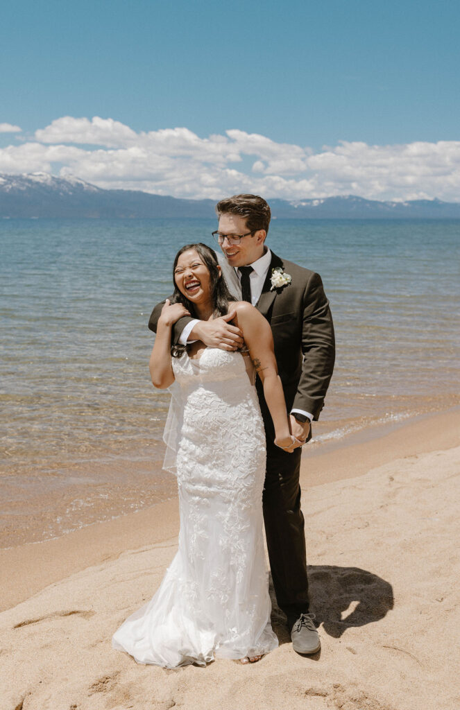 Elopement groom hugging bride while she holds his arm and both laugh on Lake Tahoe beach