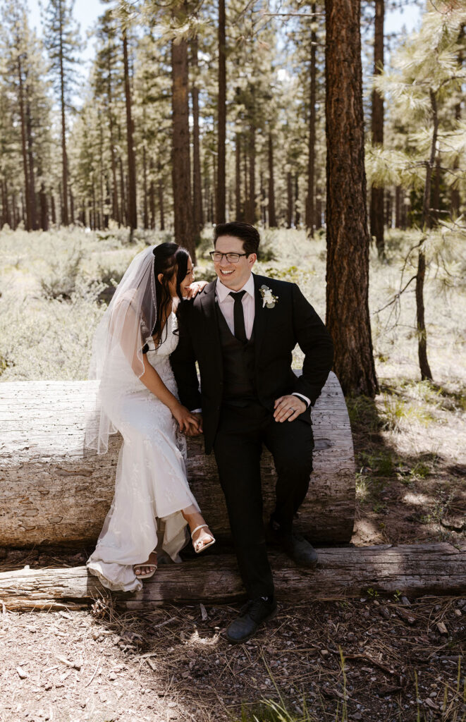 Elopement couple sitting on wooden log together while groom smiles with tall pine trees in background in Lake Tahoe