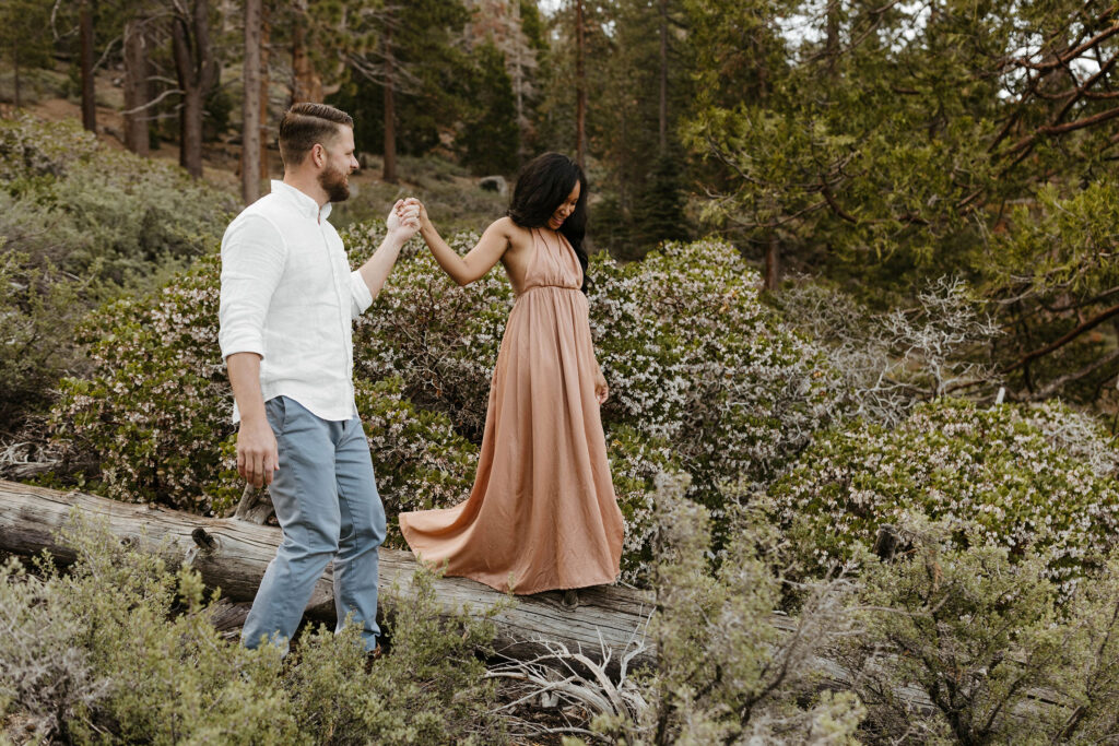 Husband holding wife's hand as she walks along log with bushes all around in Lake Tahoe