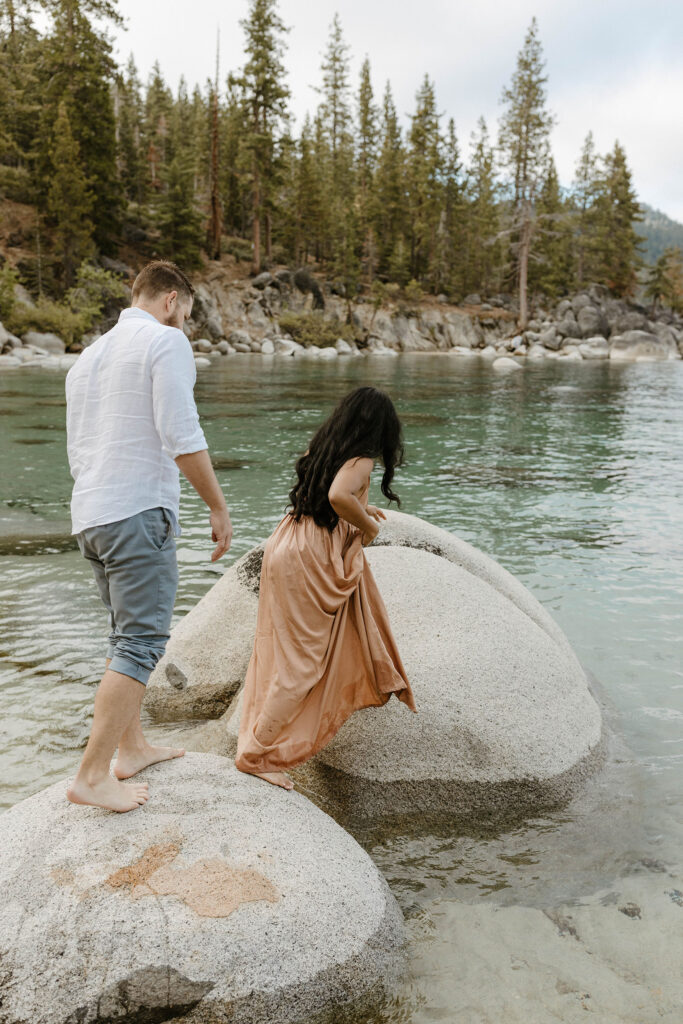 Married couple barefoot and walking across large rocks in water at Lake Tahoe with tall trees in background