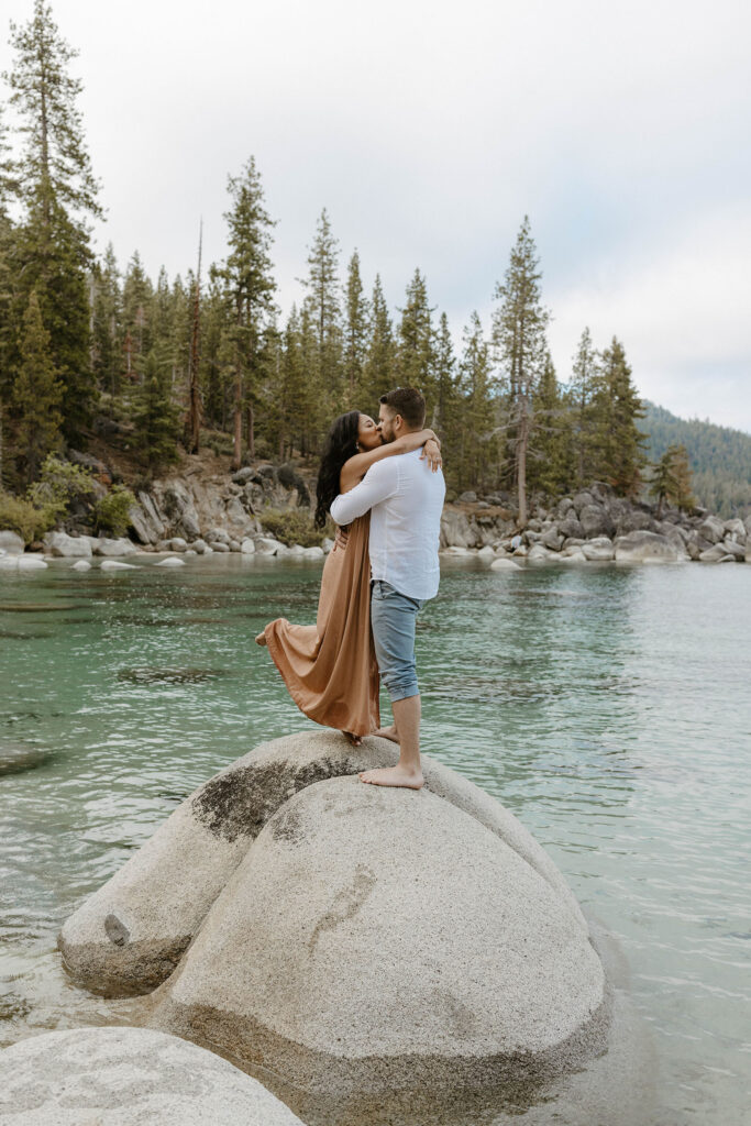 Married couple standing on large rock in Lake Tahoe while kissing with pine trees in background