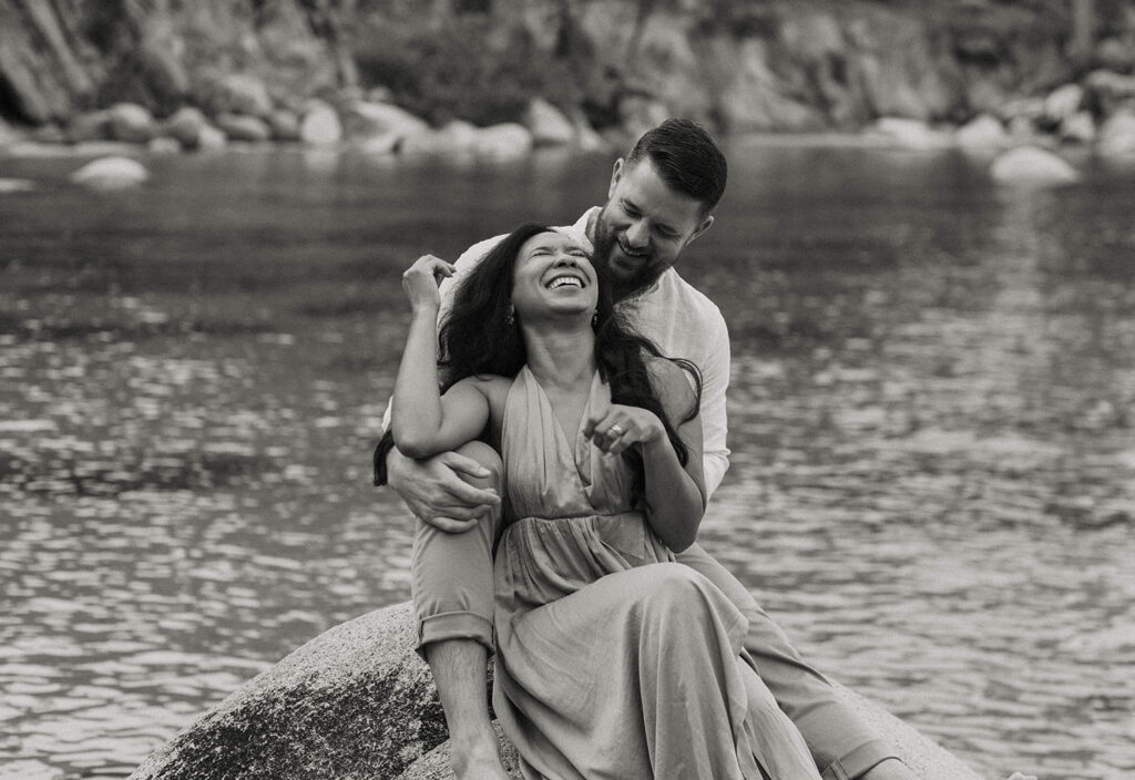 Husband holding wife as they both sit on large rock together laughing with Lake Tahoe in background