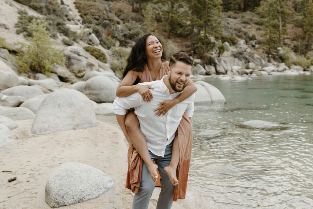 Husband giving wife piggyback ride while on a sandy beach in Lake Tahoe with rocks and trees in background