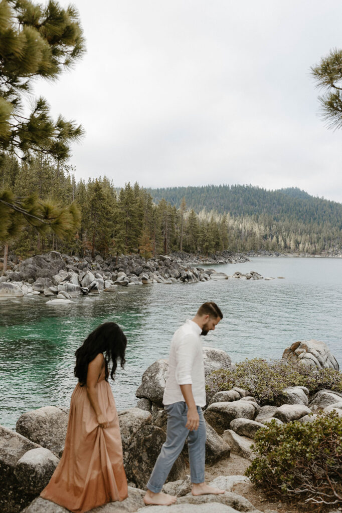 Married couple walking along rock trail barefoot with lots of rocks and pine trees in background at Lake Tahoe