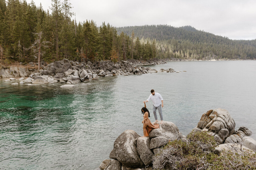 Husband holding wife's hand as she walks down large rocks with Lake Tahoe in background 
