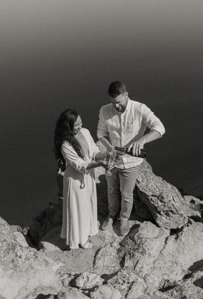 Married couple pouring champagne into champagne glasses while standing on rocks high above Lake Tahoe with lake below