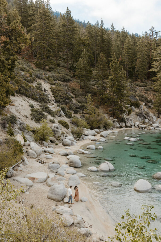 Far away shot of married couple on sandy beach in Lake Tahoe with lots of rocks
