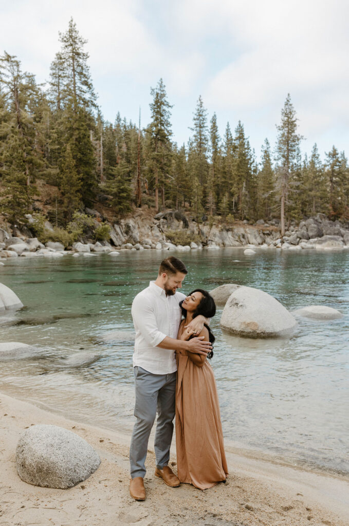 Married couple looking at each other while smiling on Lake Tahoe beach with large rocks and trees in background