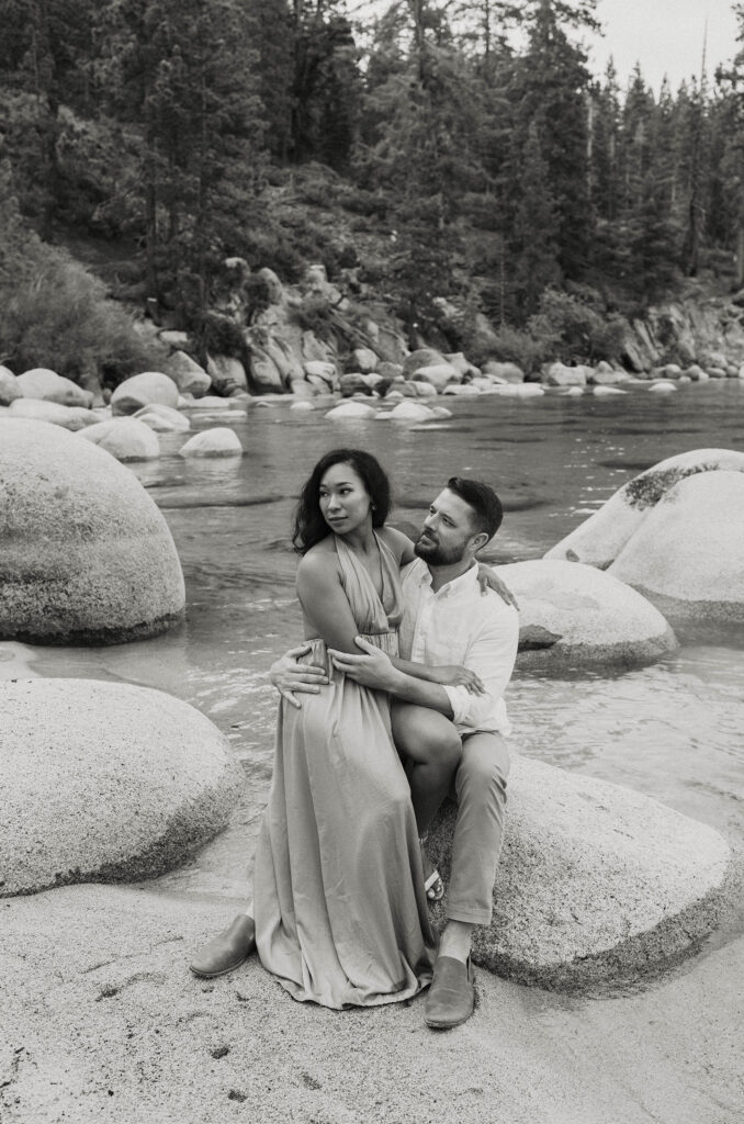 Husband sitting on rock while wife sits on his leg and they look into distance together on beach in Lake Tahoe