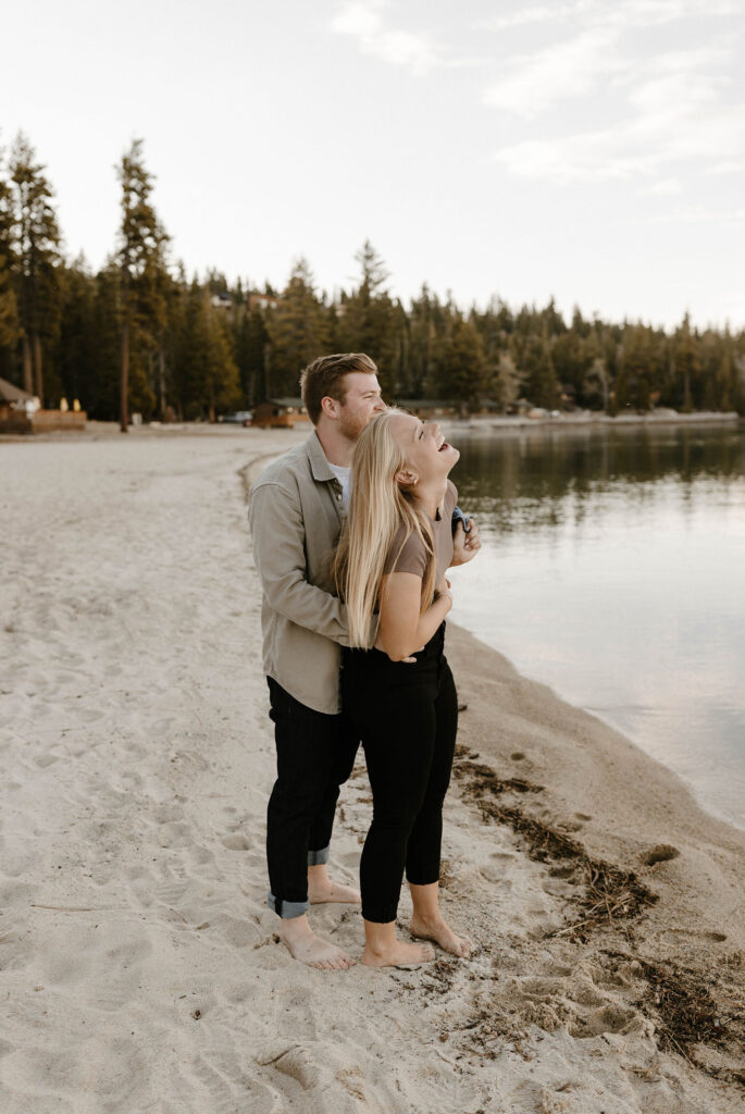 Woman laughing while fiancé is hugging her from behind standing on a beach together at Meeks Bay