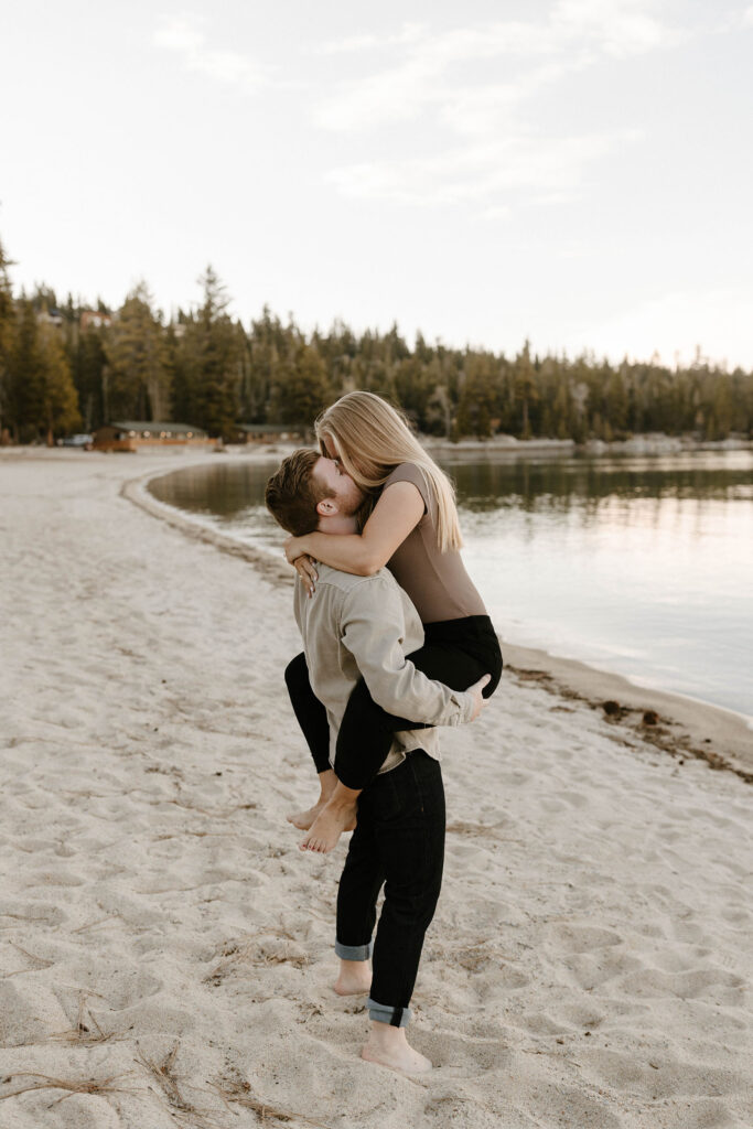 Man picking up fiancé and kissing her while on sandy beach at Meeks bay with tall trees and water in background