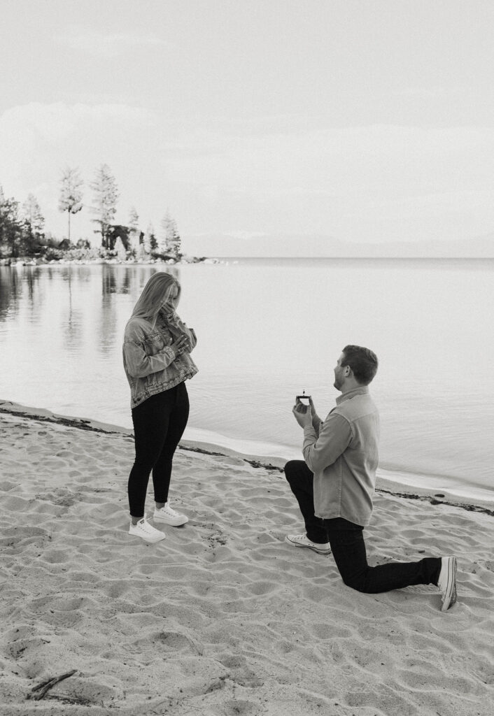 Woman emotional while fiancé is on one knee holding engagement ring while on sandy beach at Meeks Bay