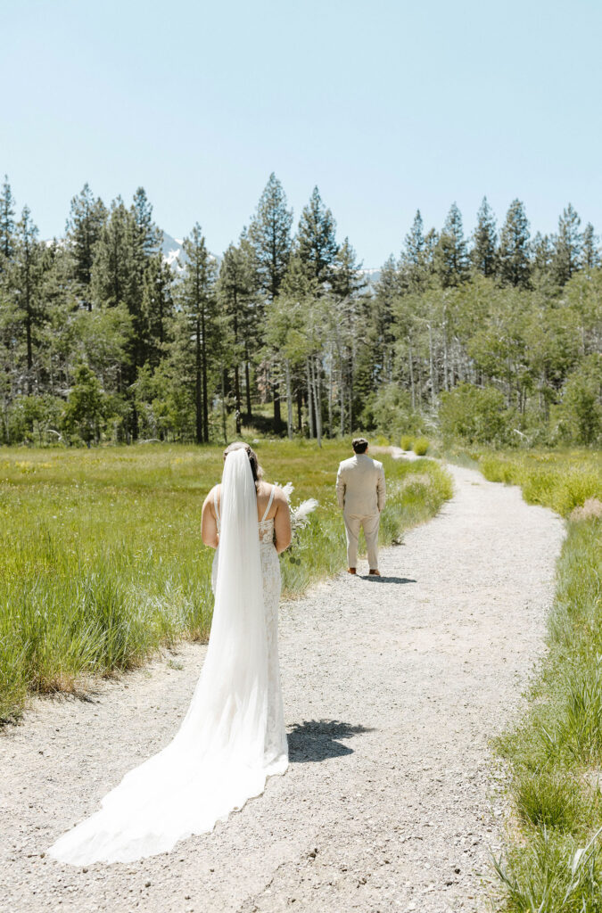 Wedding bride holding floral bouquet while walking up to groom facing away during first look on dirt trail next to grassy meadow in Lake Tahoe