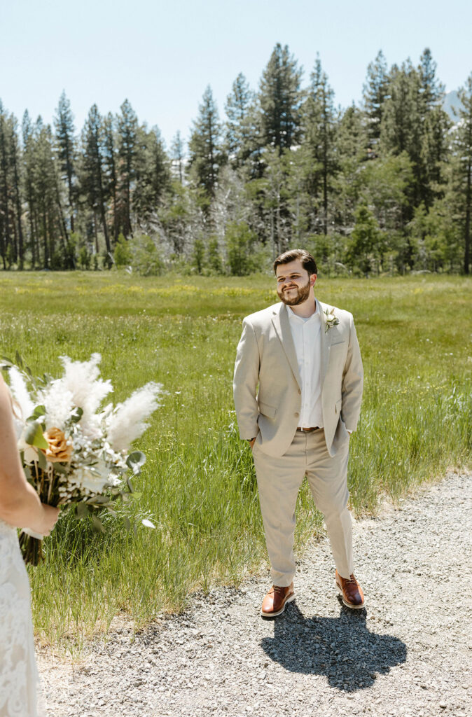Wedding groom smiling while turning around to see bride during first look while on dirt trail with grass meadow behind him in Lake Tahoe