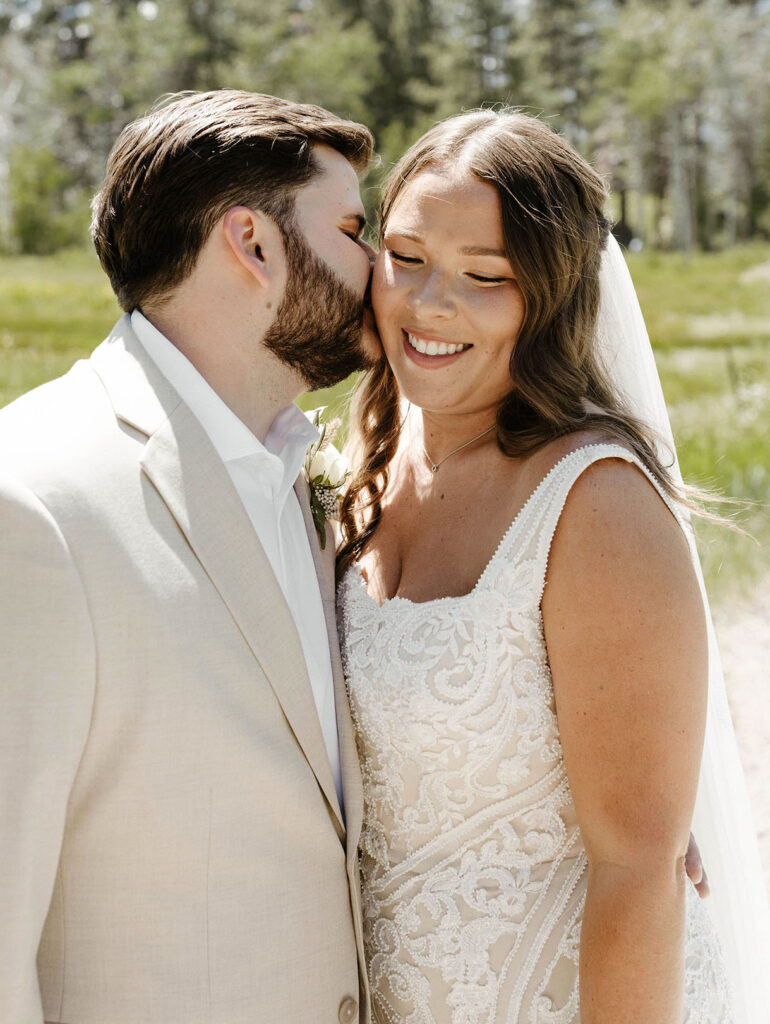 Groom kissing wedding bride on cheek while she smiles with grassy meadow behind them in Lake Tahoe