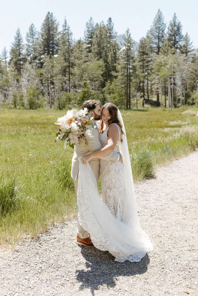 Groom holding bride and kissing her cheek while she looks down and smiles while standing on dirt trail with grassy meadow in background in Lake Tahoe