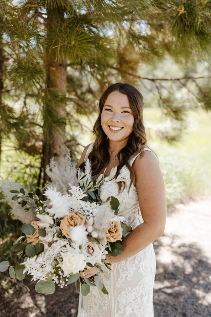 Wedding bride holding floral bouquet while smiling at camera in front of pine tree in Lake Tahoe