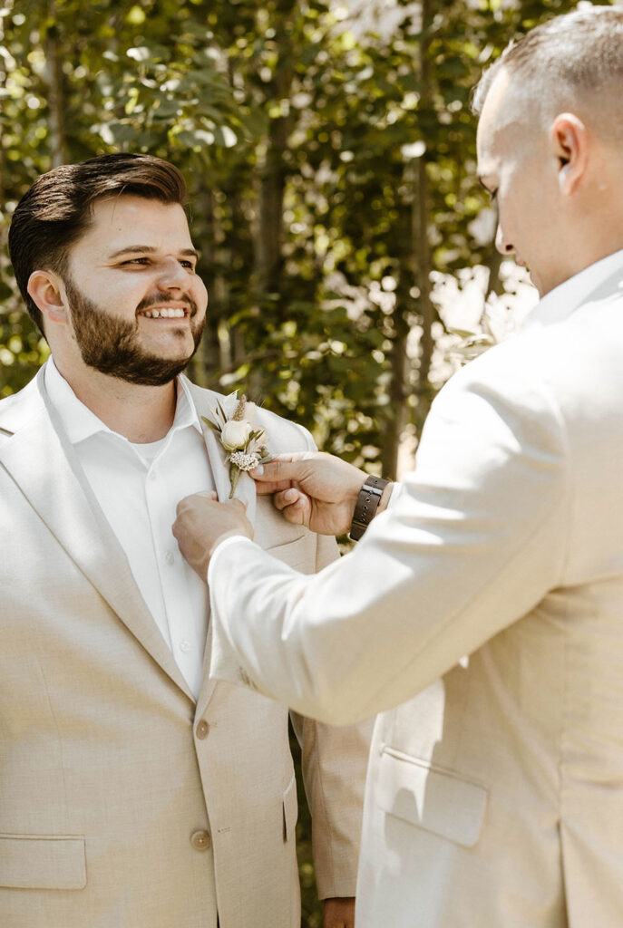 Best man adjusting Boutonnière on grooms suit while both are smiling outside at Lake Tahoe