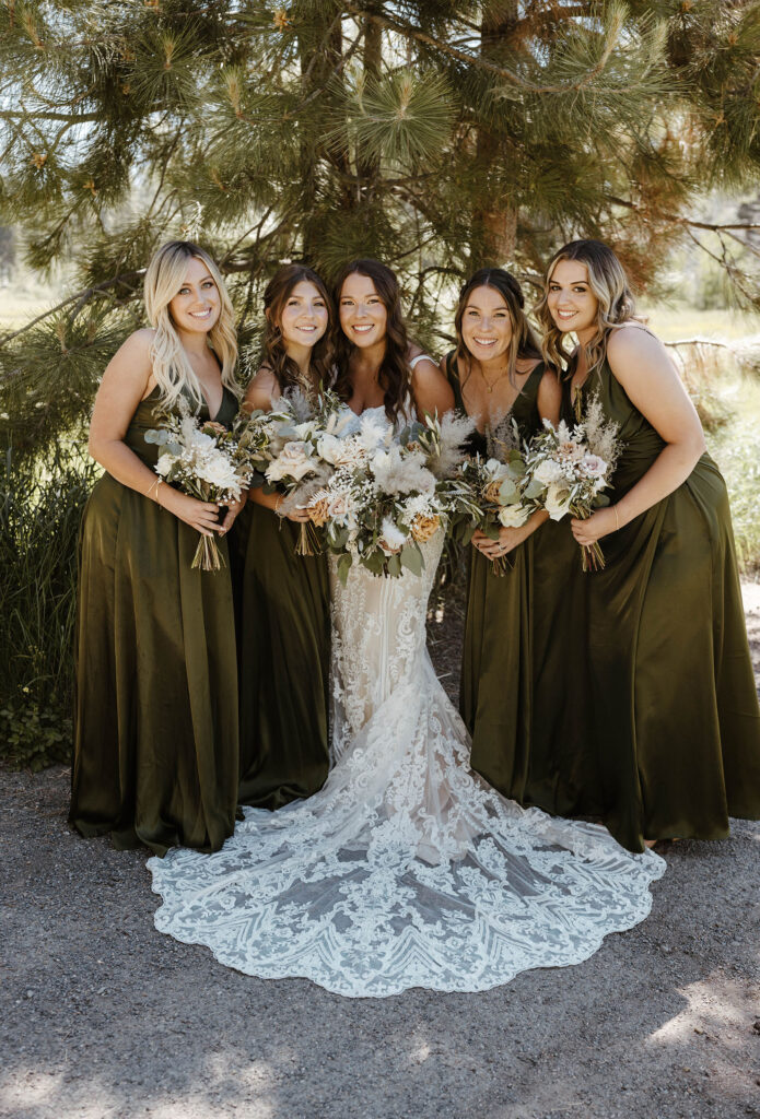 Wedding bride and bridesmaids standing close together and smiling at the camera in front of pine tree in Lake Tahoe