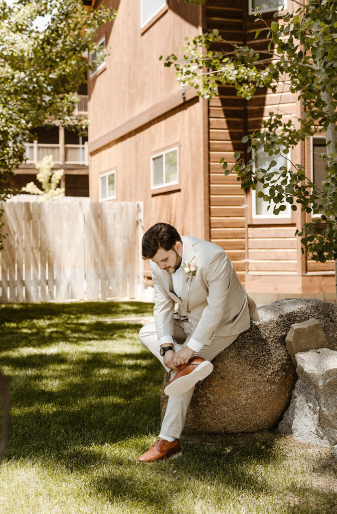 Groom sitting on large rock while tying wedding shoes with wooden house and fence in background in Lake Tahoe