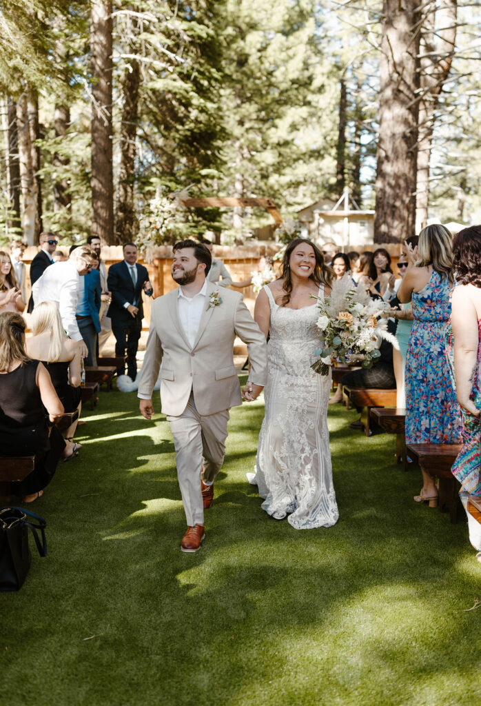 Wedding couple holding hands and smiling at guests while walking down aisle together after ceremony in Lake Tahoe