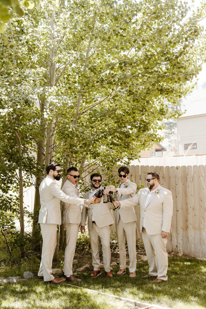 Groom and groomsmen cheering beer bottles while standing together outside next to a fence and tree in Lake Tahoe