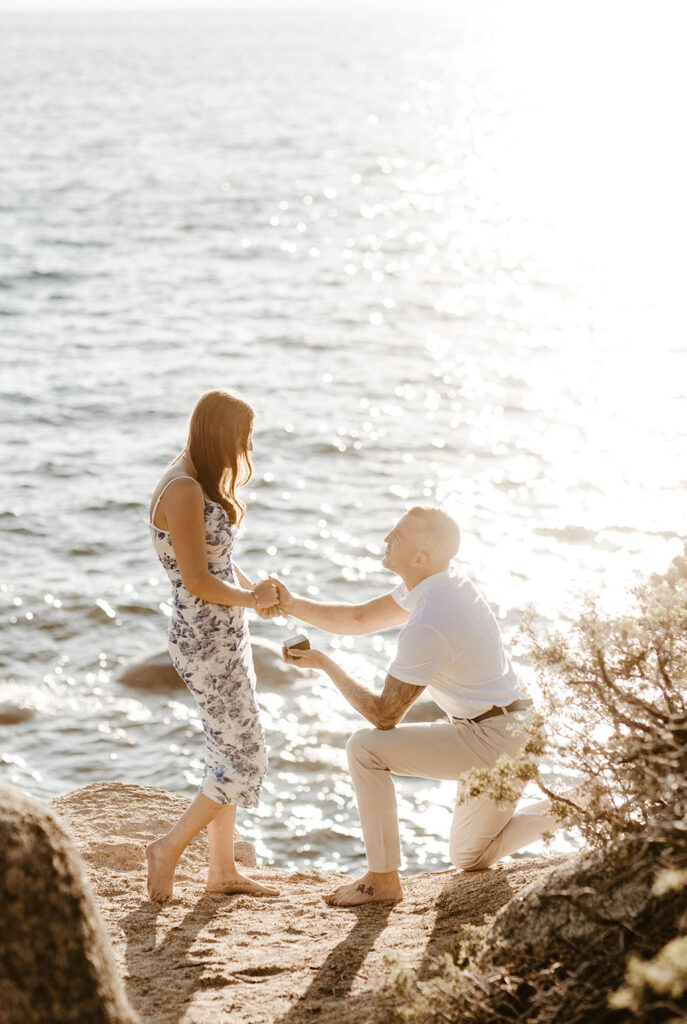 Man on one knee holding woman's hand and proposing while on sand next to Lake Tahoe