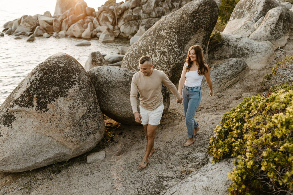Man leading fiancé down rock and sand beach barefoot while both smile at Lake Tahoe