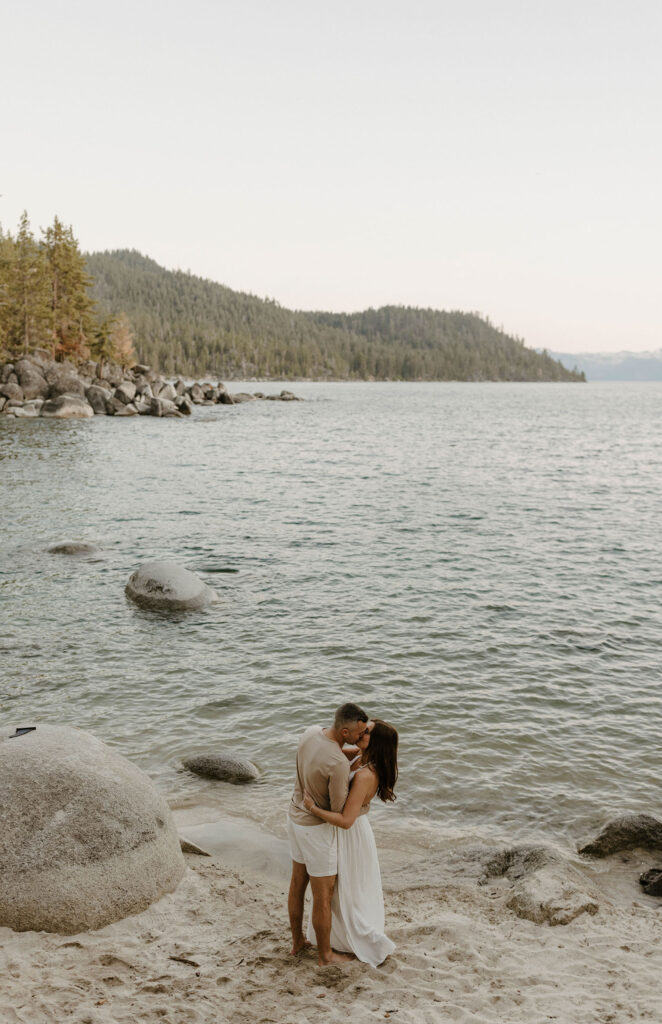 Engagement couple holding each other as they kiss on sandy beach with Lake Tahoe and pine trees in background