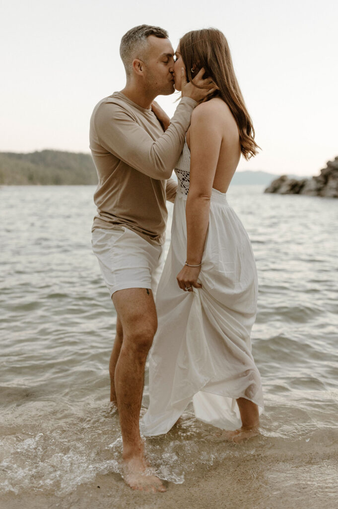 Engagement couple kissing on sandy beach while water crashes over their feet at Lake Tahoe