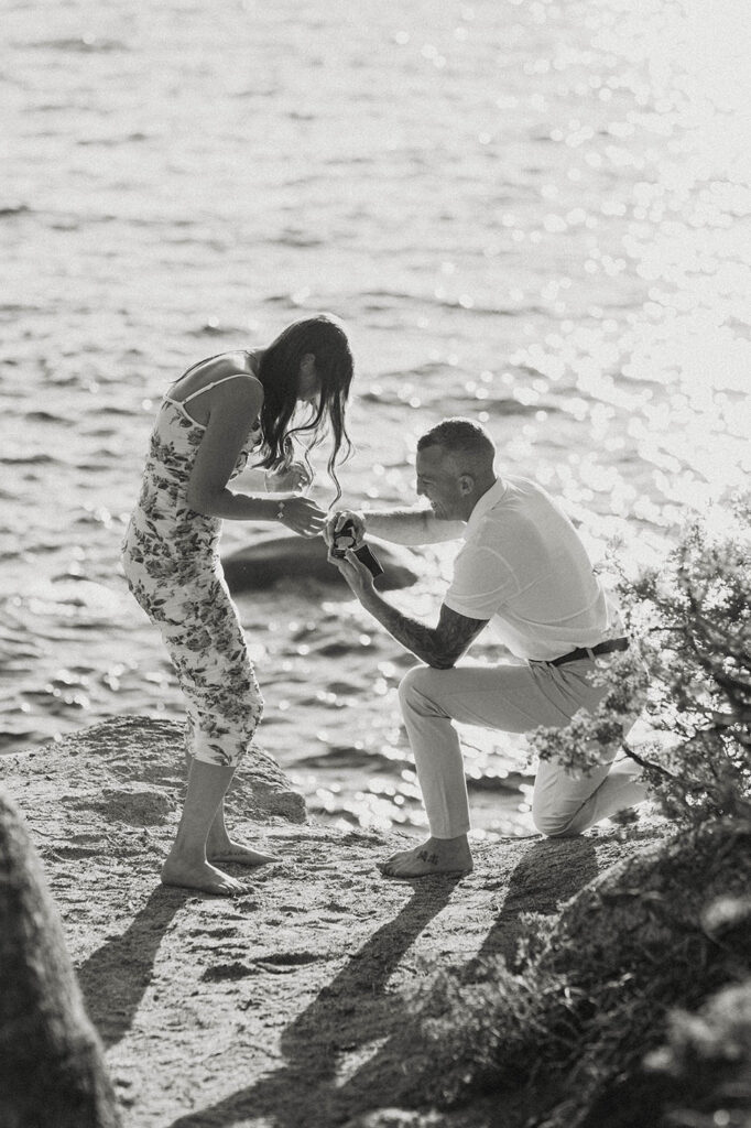 Man putting ring on fiancé as both laugh while on sand with Lake Tahoe in background
