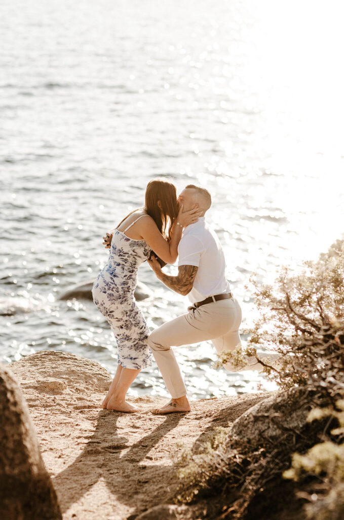 Woman excitedly kissing fiancé while holding his face with both hands on sand with Lake Tahoe in background