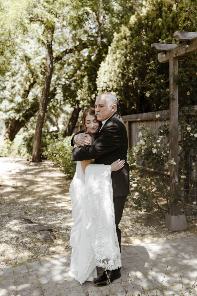 Wedding bride and dad hugging with eyes closed after first look on stone path with trees in background at French Oak Ranch