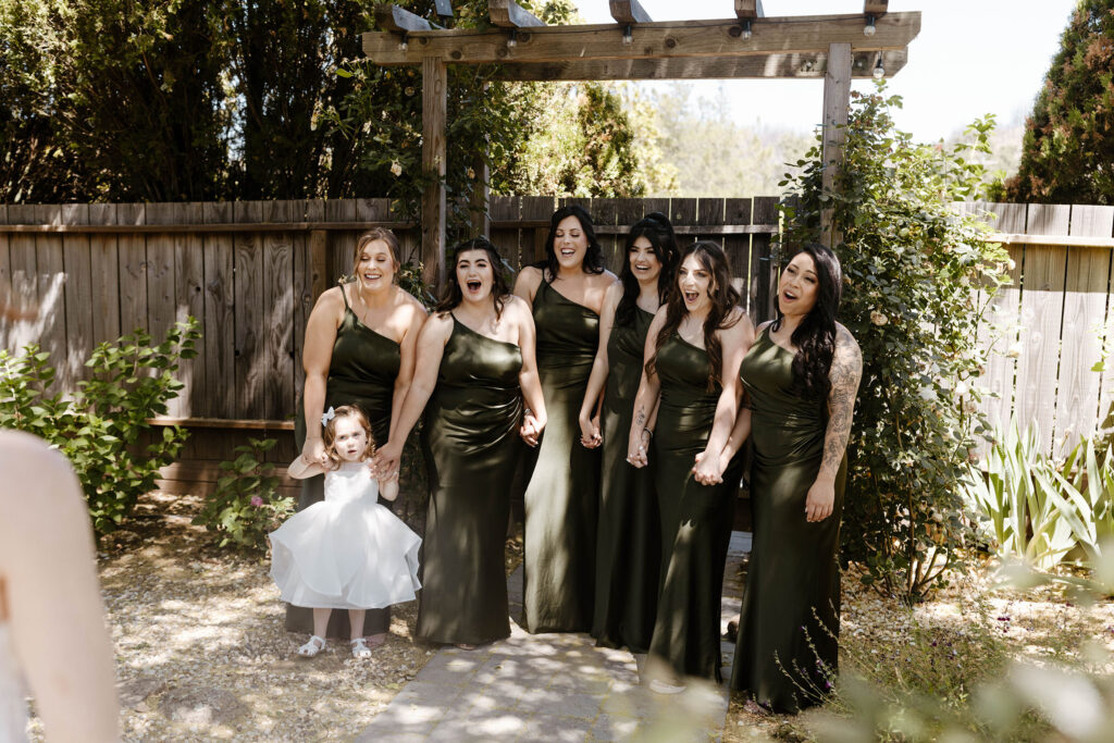 Bridesmaids reacting excited and happy during first look with bride and wedding dress while holding hands in front of wooden fence at French Oak Ranch