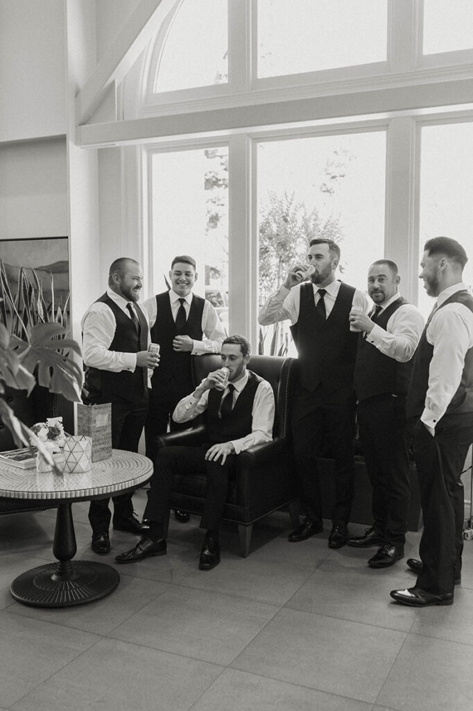 Wedding groom sitting in leather chair drinking while groomsmen stand around him with beers at French Oak Ranch