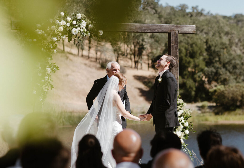 Wedding couple holding hands during wedding ceremony while groom laughs with wooden arch and white florals behind them at French Oak Ranch