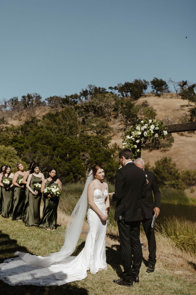Bride looking at groom reading vows during ceremony at French Oak Ranch with bridesmaids and hills in background