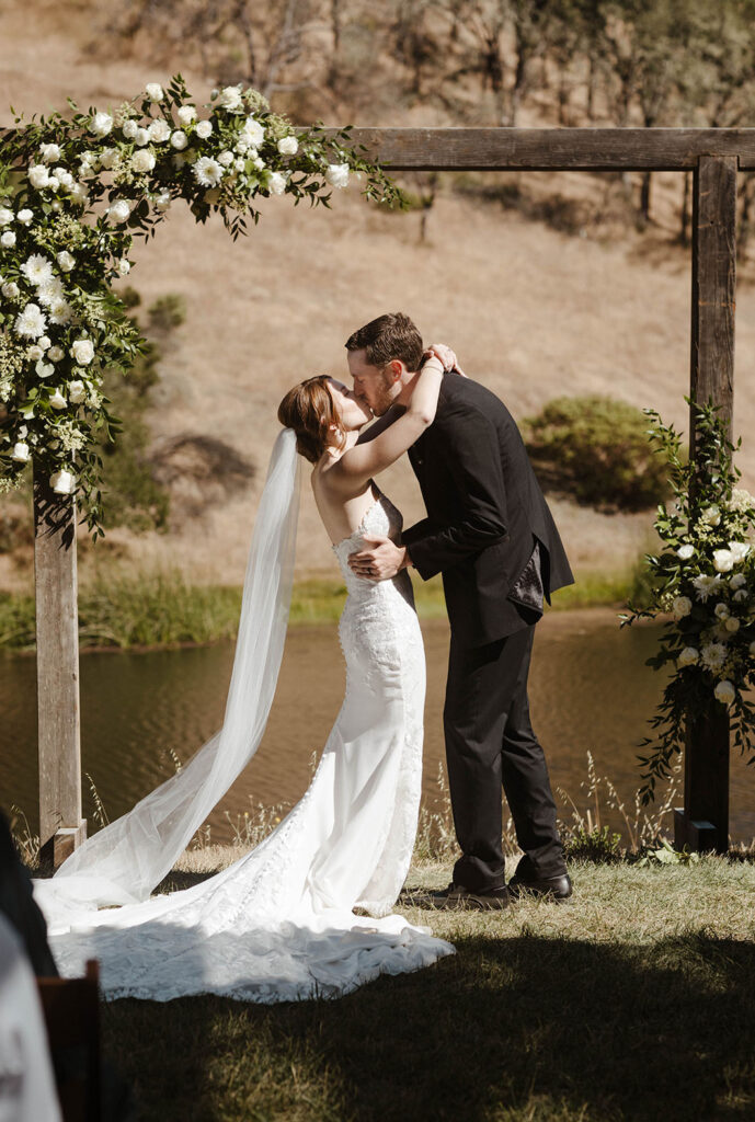 Wedding couple's first kiss inside wooden arch with white florals during ceremony at French Oak Ranch